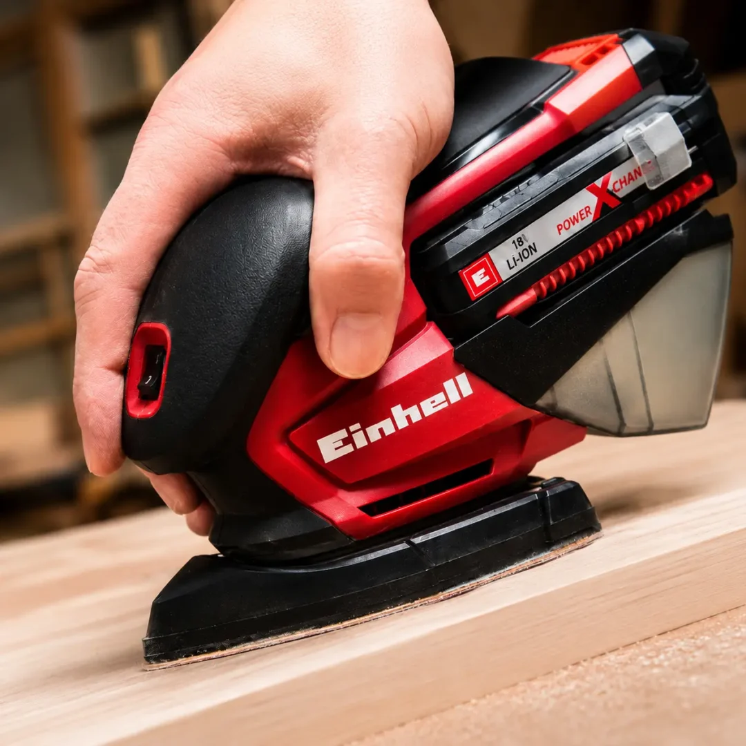 Lijadora Einhell roja y negra a batería trabajando sobre una tabla de madera, con mano sujetando la herramienta, depósito de polvo y sistema Power X-Change visibles.